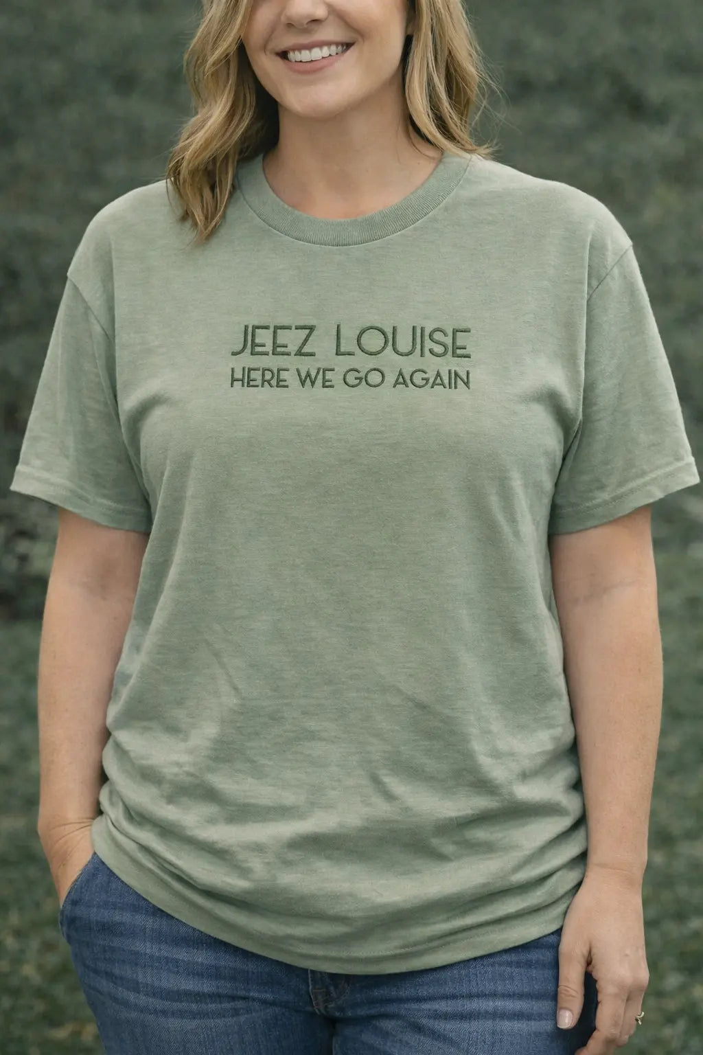 Woman wearing a green t-shirt with the phrase "JEEZ LOUISE HERE WE GO AGAIN" printed in dark green.