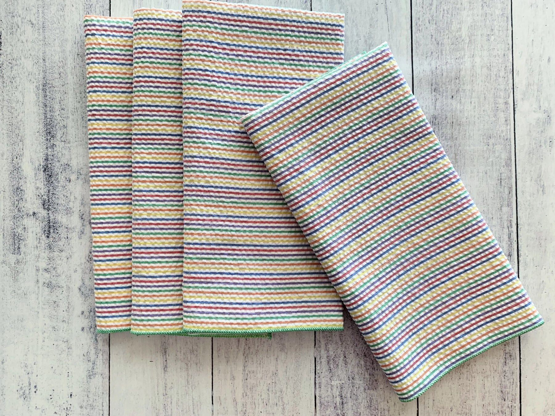 Set of four rainbow stripe seersucker cloth napkins stacked on a white wood background.