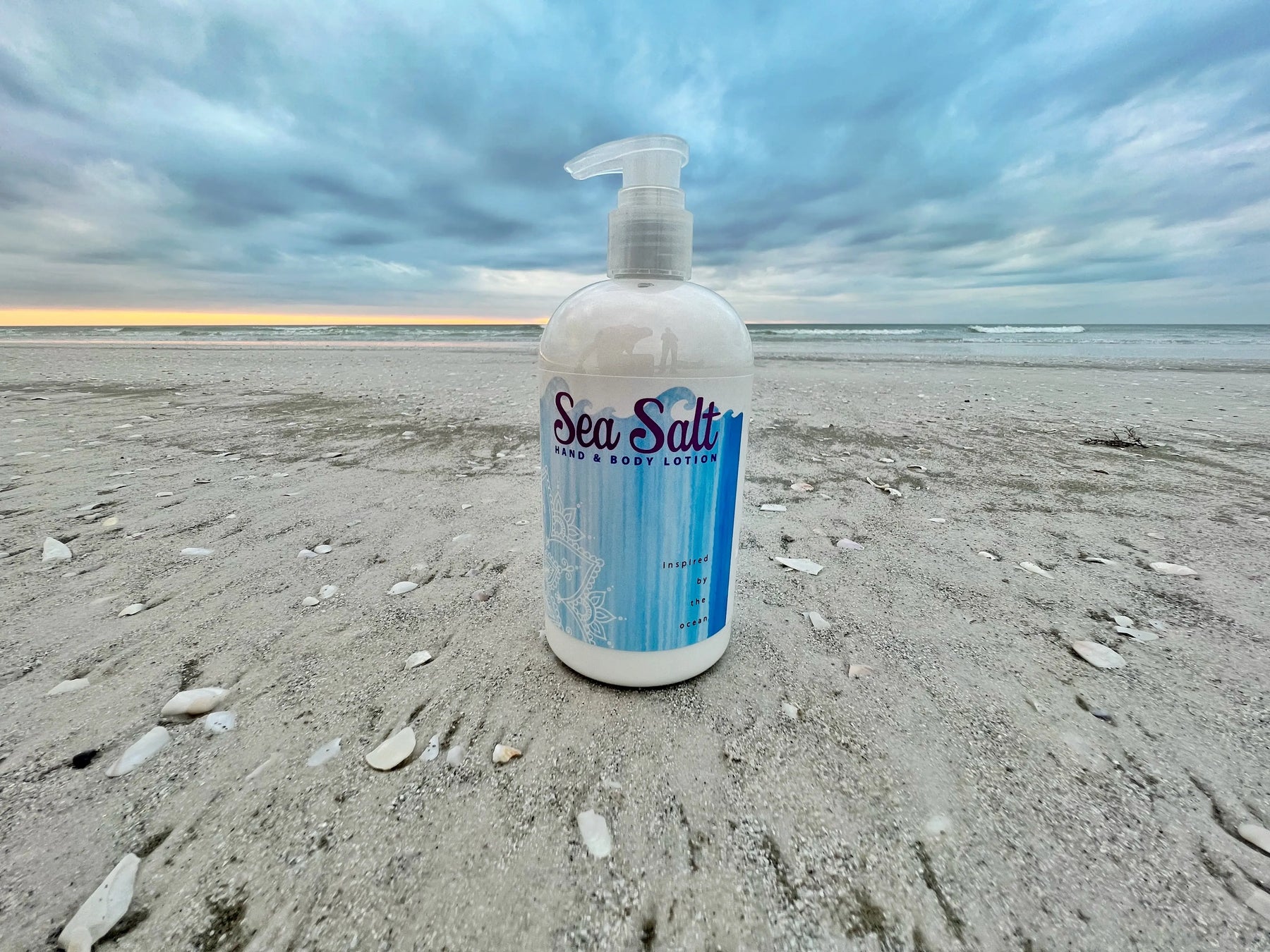 Sea Salt Hand & Body Lotion bottle on a sandy beach with the ocean and cloudy sky.