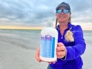 Woman holding a bottle of Sea Salt Hand & Body Lotion on a beach with the ocean in the background.