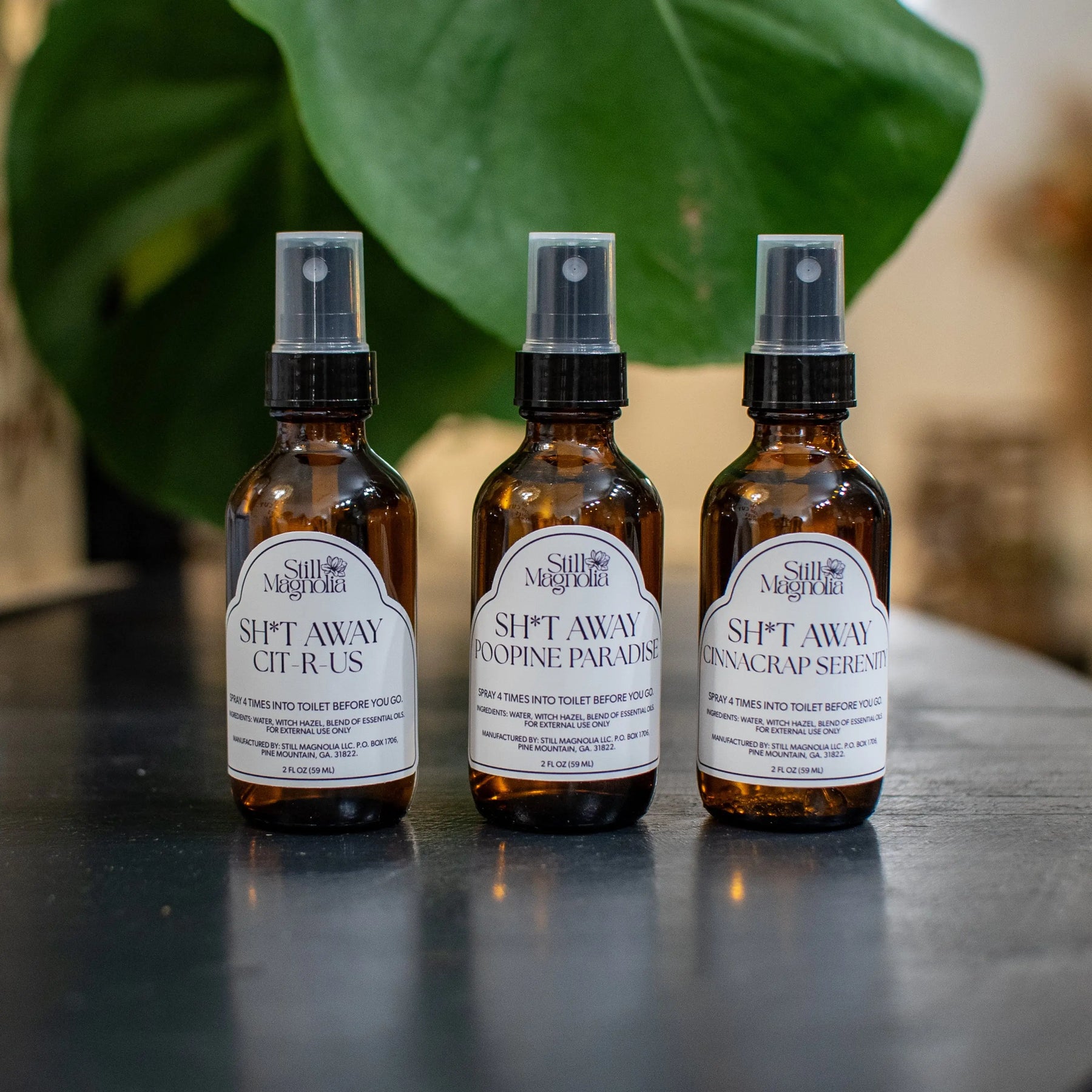 Three amber glass bottles of Still Magnolia Sh*t Away bathroom spray on a dark surface.