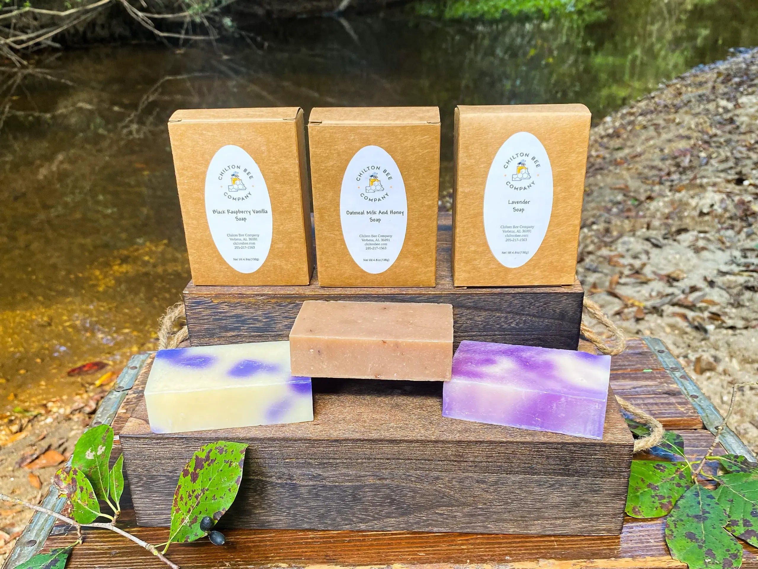 Three Chilton Bee Company soap boxes with soap bars displayed on a wooden stand in front of a stream.
