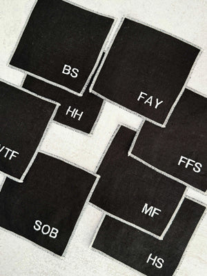 Black linen coasters with white embroidered initials arranged on a surface.