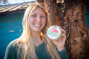 Woman holding a round tin of grapefruit and calendula hand cream next to a tree trunk