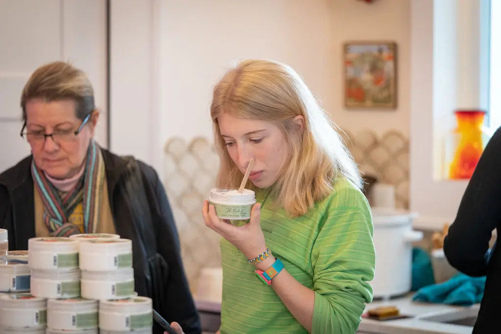 Young woman smelling jar of "The Orchard" grapefruit and calendula hand cream with wooden applicator.