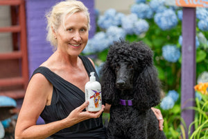 Woman holds a bottle of goat milk lotion next to a black poodle outdoors.