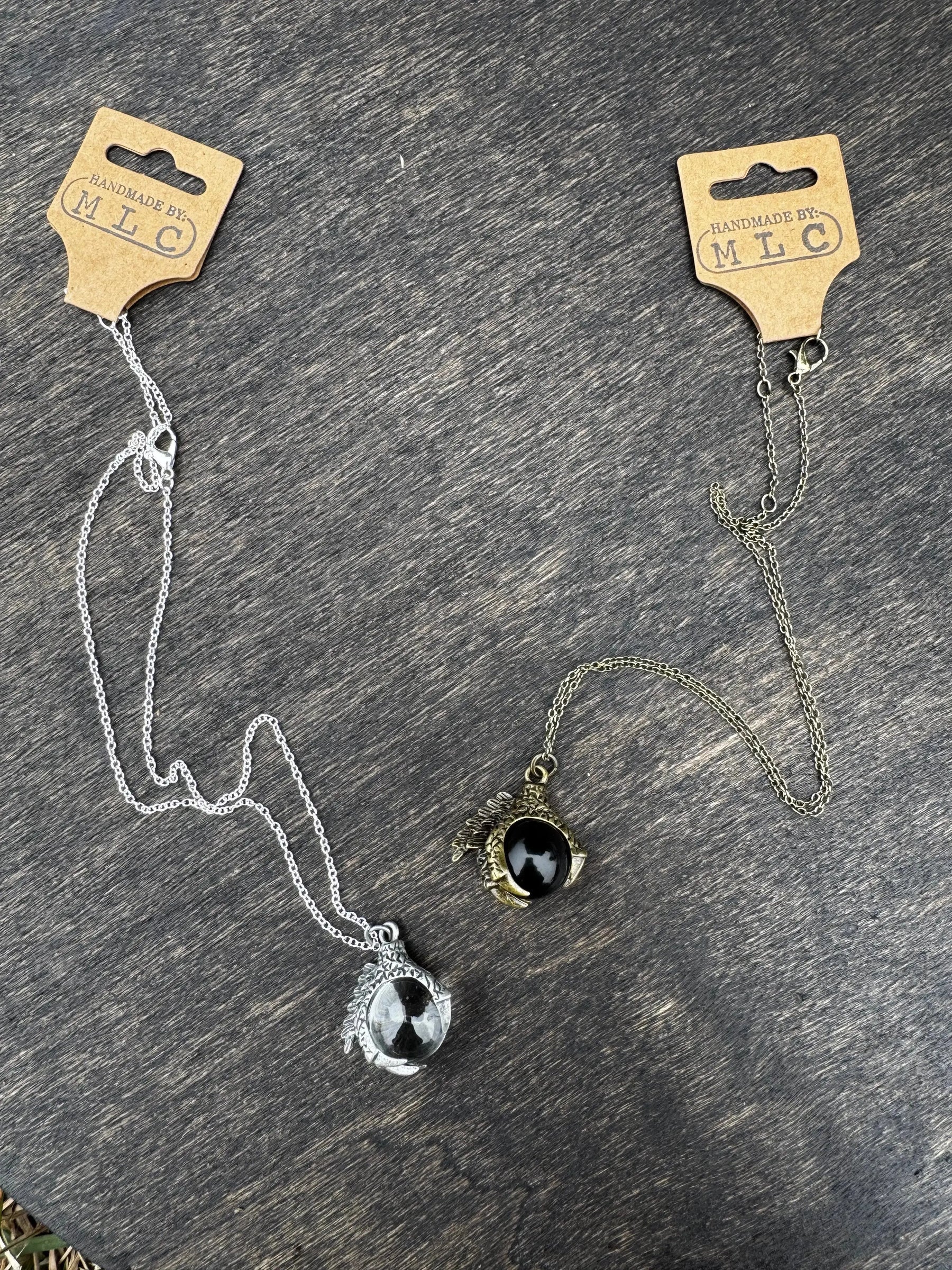 Two dragon claw necklaces on a wood surface. One silver with a clear orb, the other gold with a black orb.
