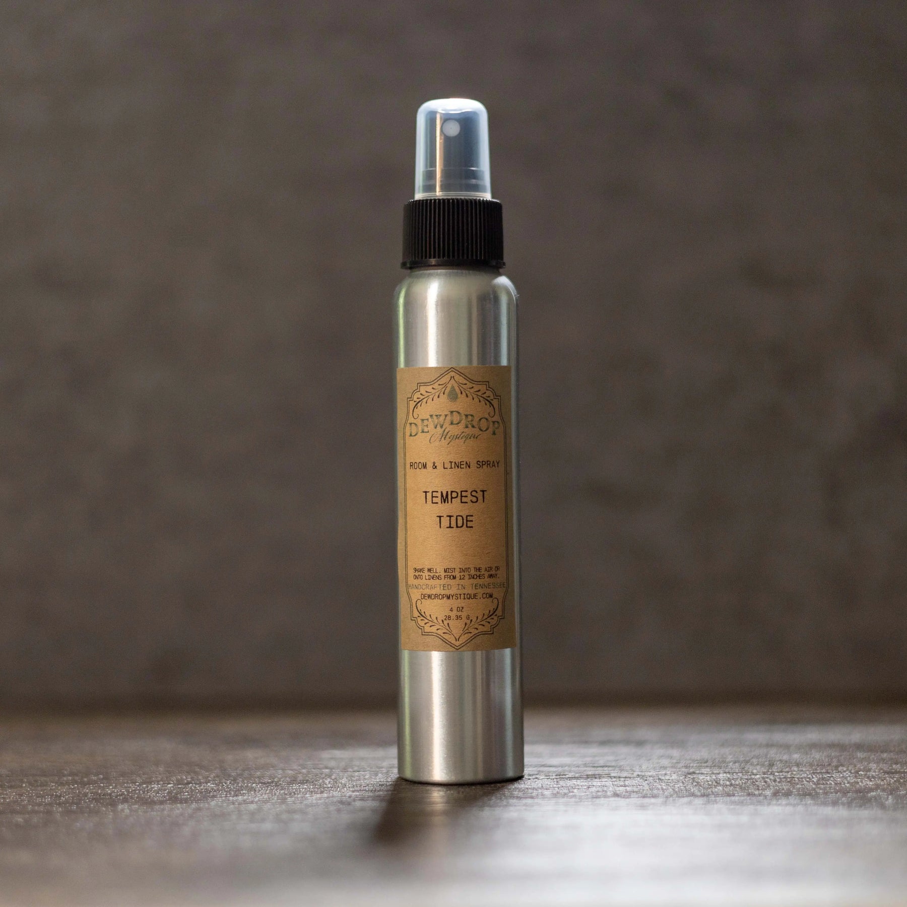 Silver spray bottle with a brown label reading "Tempest Tide Room & Linen Spray" on a dark wooden surface.