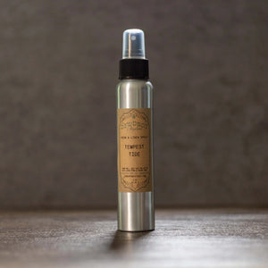 Silver spray bottle with a brown label reading "Tempest Tide Room & Linen Spray" on a dark wooden surface.