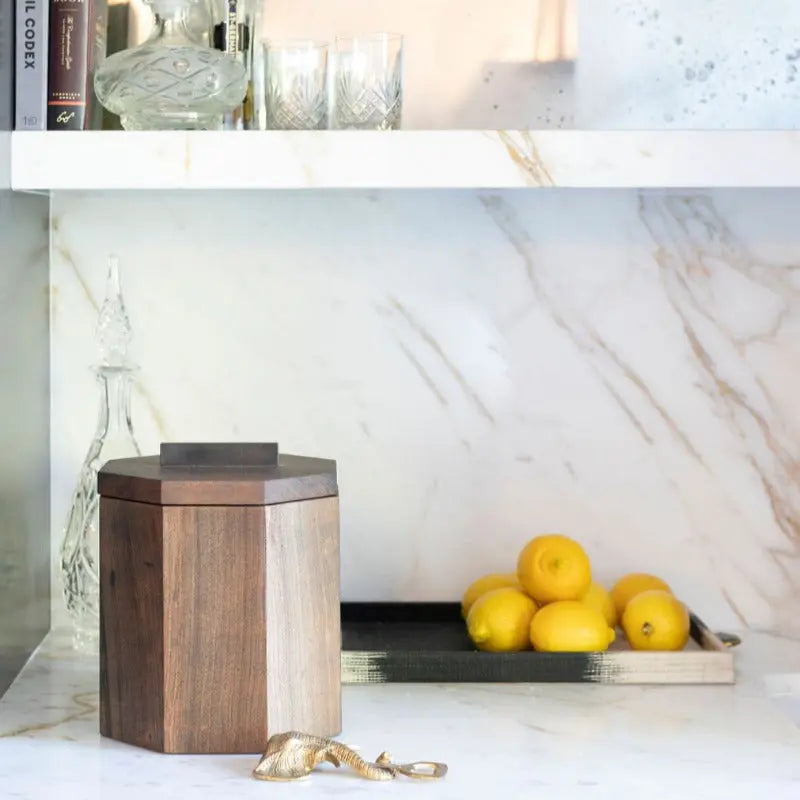 Wooden ice bucket with blackened cherry lid, lemons, and a gold bottle opener on a marble countertop.