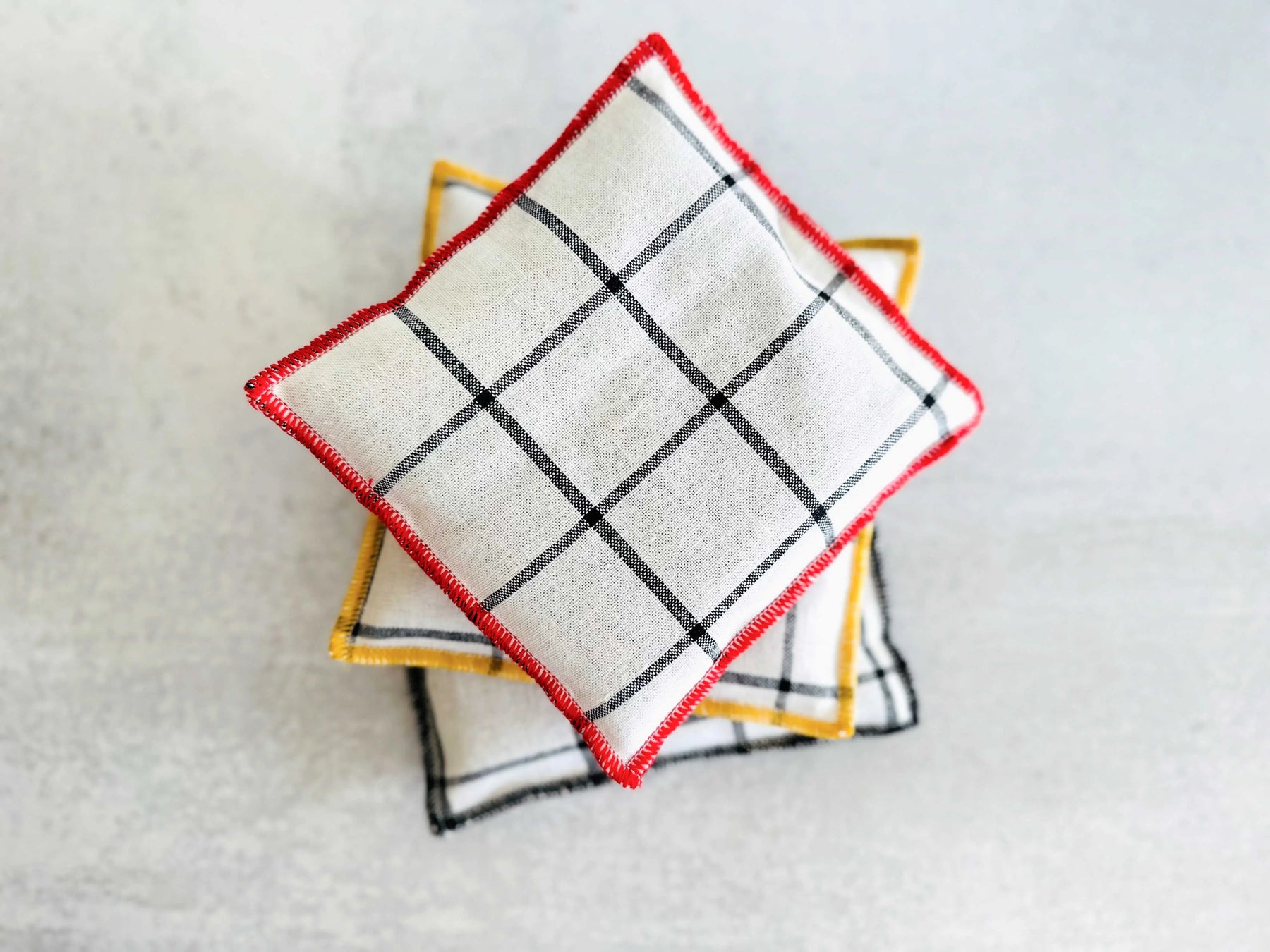 White linen sachets with black plaid pattern and colored edges stacked on a gray surface.