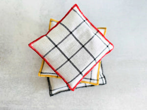 White linen sachets with black plaid pattern and colored edges stacked on a gray surface.