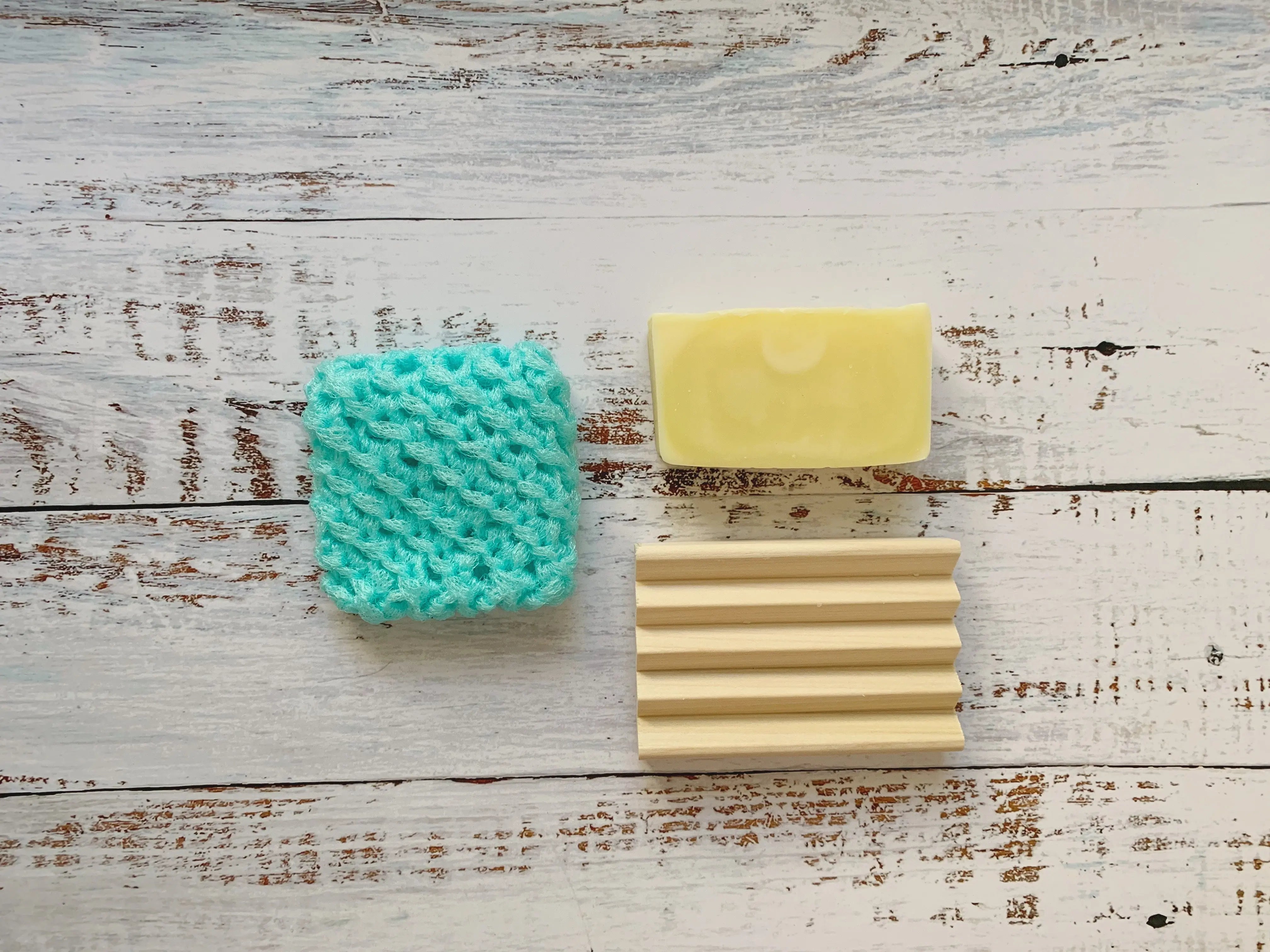Light yellow bar soap, textured teal sponge, and ribbed wooden soap dish on white wood background