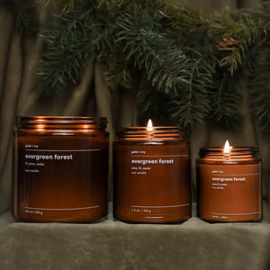 Three amber glass soy candles labeled 'Evergreen Forest' with pine needles and a velvet cloth background.