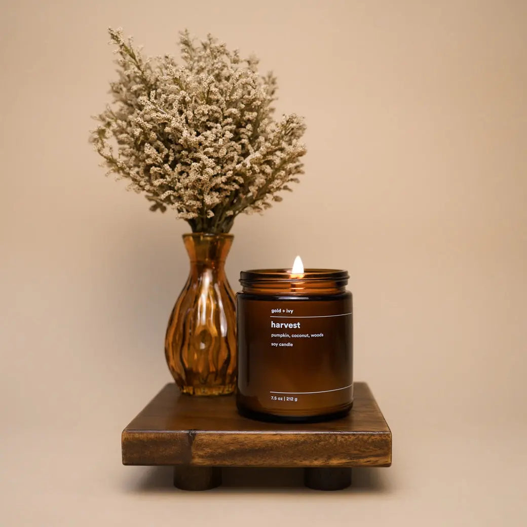 Lit harvest soy candle in amber jar with gold + ivy label on a wooden stand next to dried flowers.