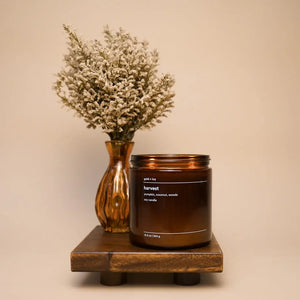 Harvest scented candle in amber glass jar with dried flowers and wooden stand.